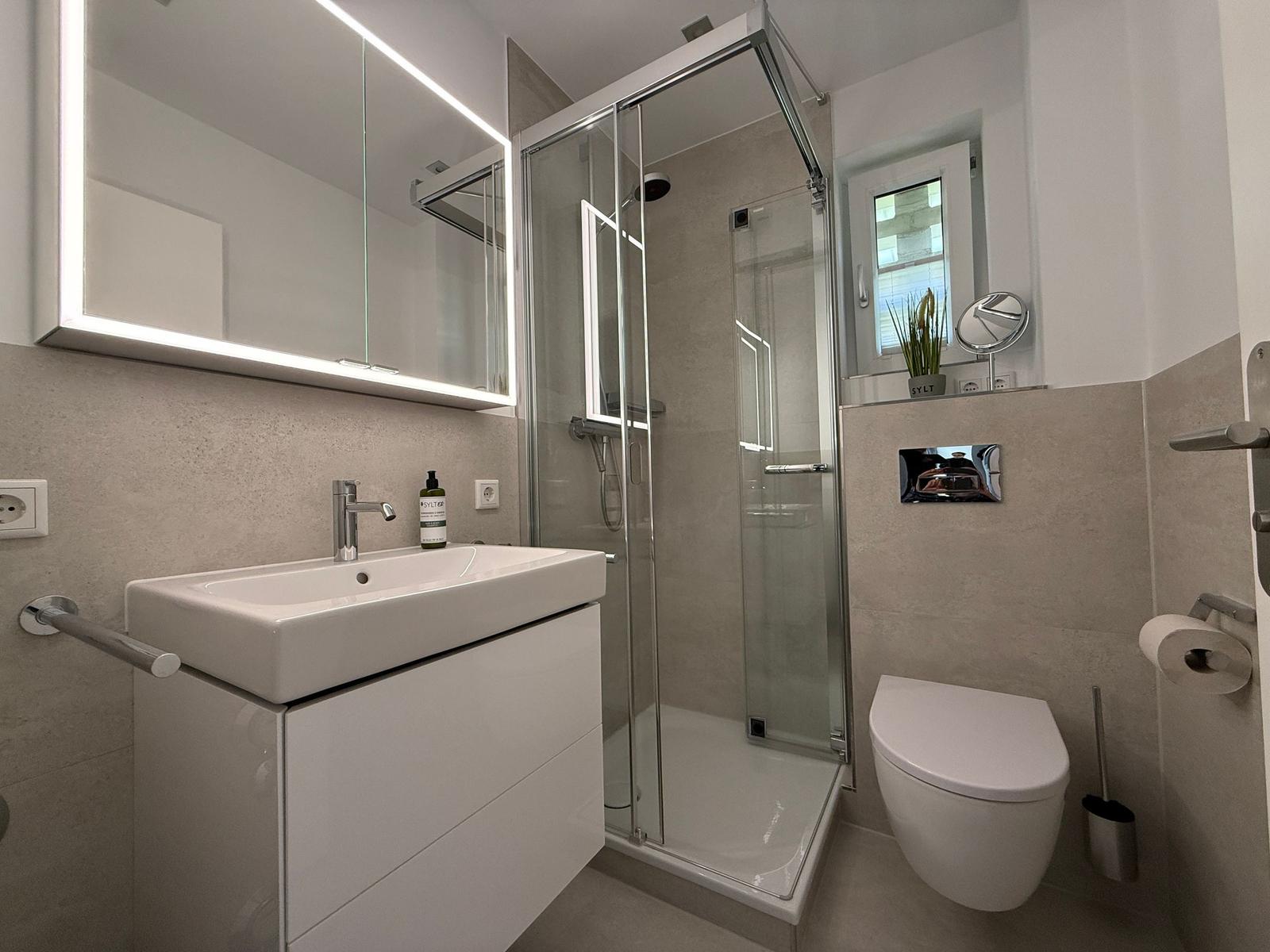 Badezimmer mit Dusche, Waschbecken und WC. Spiegel mit Beleuchtung und Fenster.