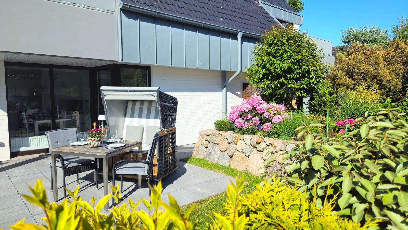 Terrasse mit Tisch, Stühlen und Liege unter blauem Himmel. Blühender Garten mit Steinmauer.