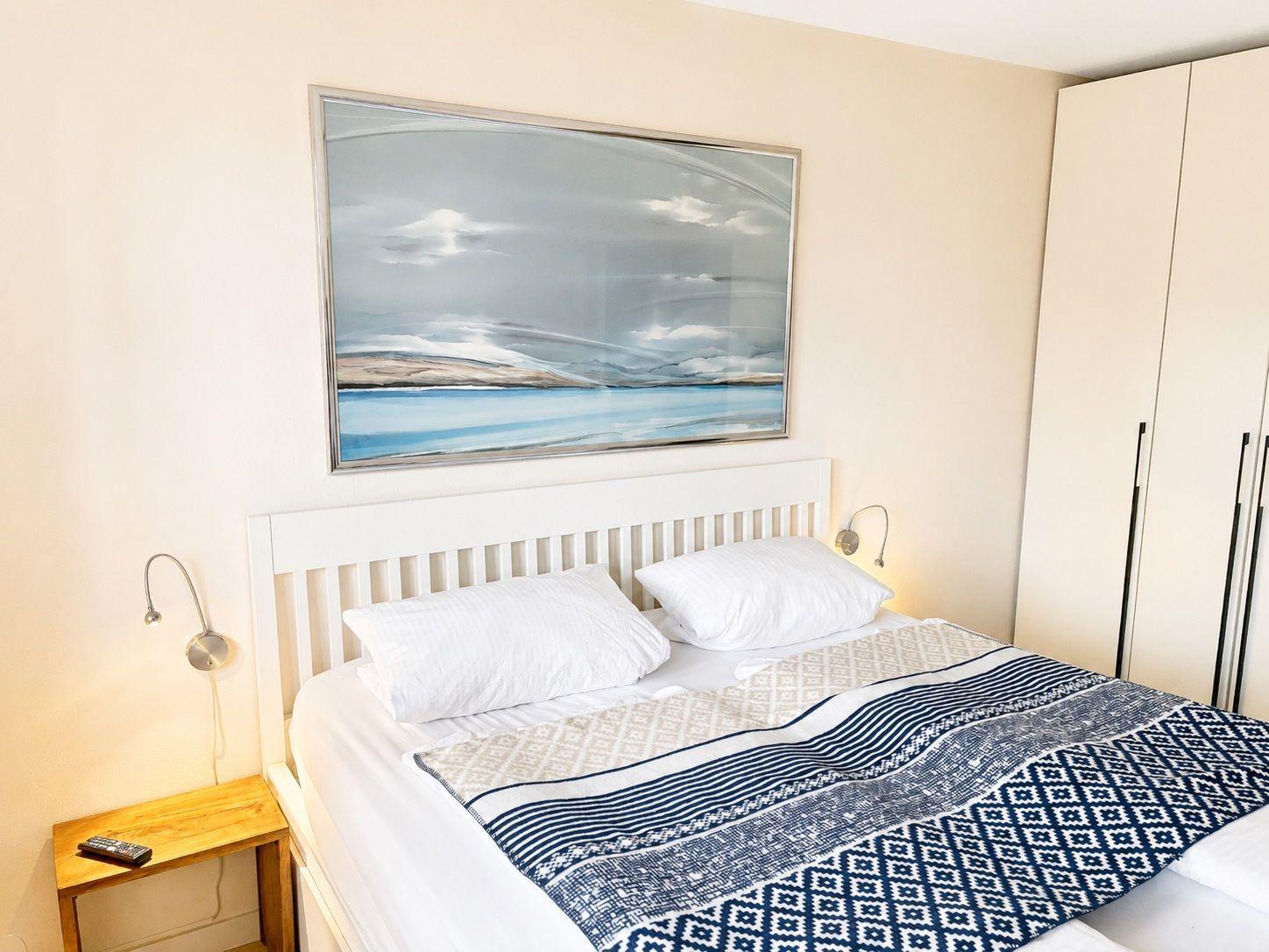 Schlafzimmer mit Bett, Kleiderschrank und Wandbild. Links Nachttisch mit Fernbedienung.