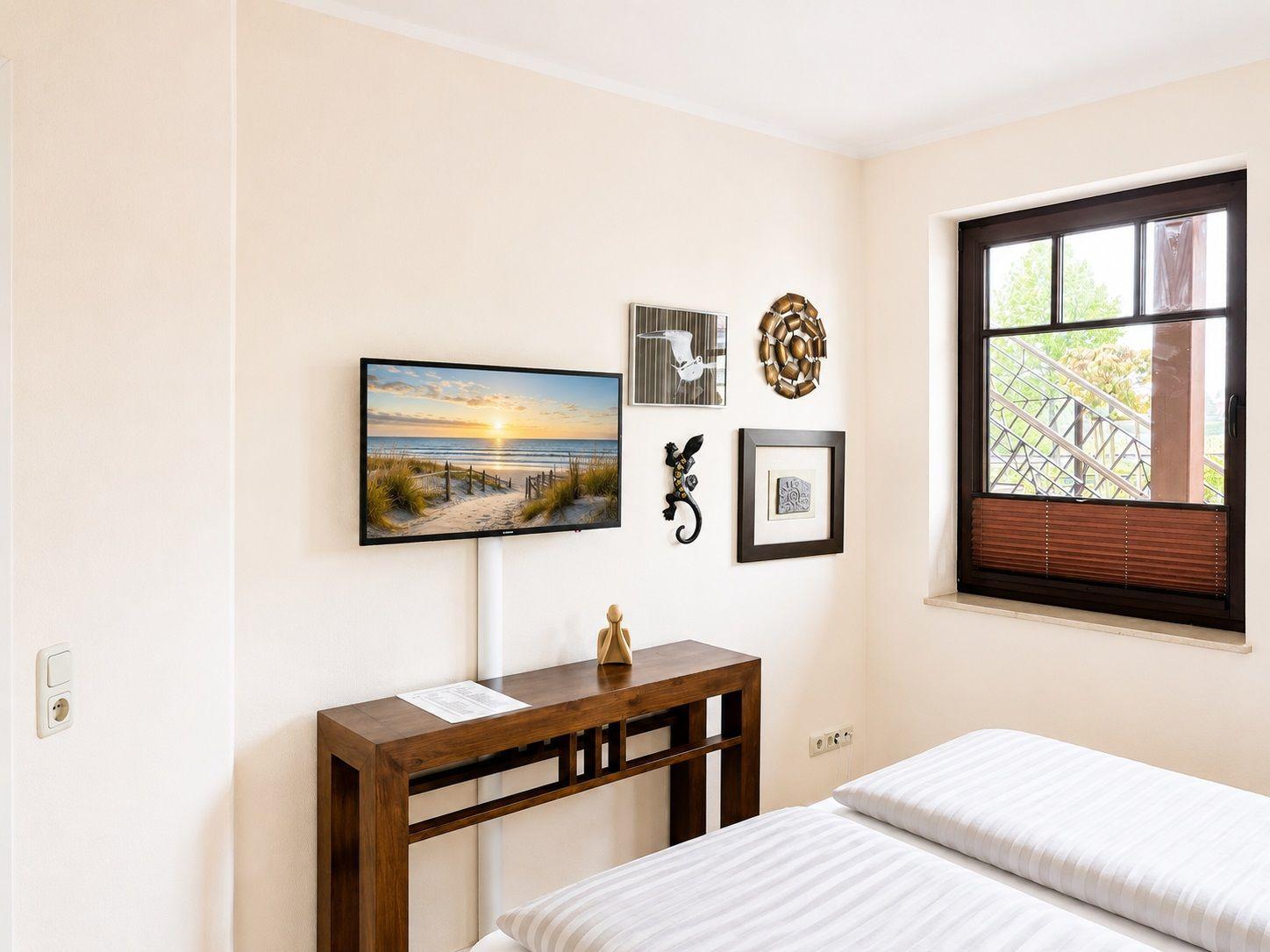 Schlafzimmer mit Bett, Wand-TV und Fenster mit Blick auf Balkon.