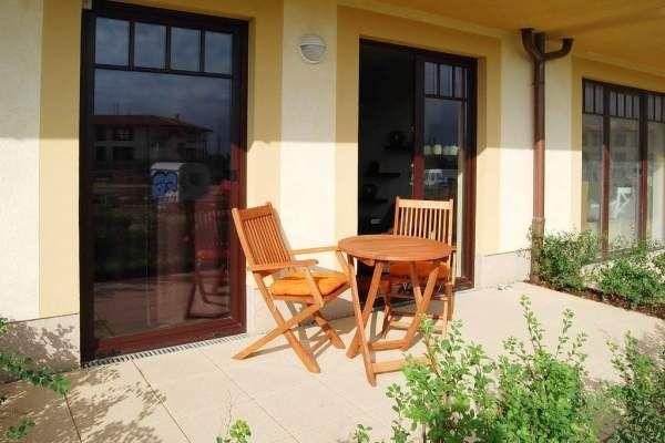 Terrasse mit Holztisch und zwei Stühlen vor einem Haus mit großen Fenstern.