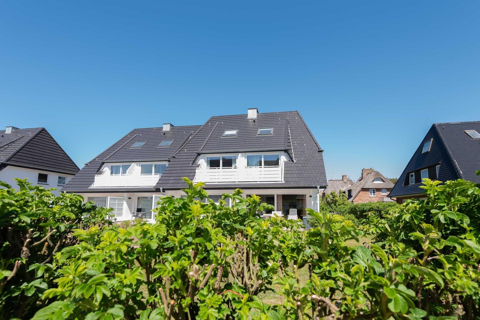 Modernes Haus mit dunklem Dach und weißen Fenstern, umgeben von grünen Sträuchern unter blauem Himmel.