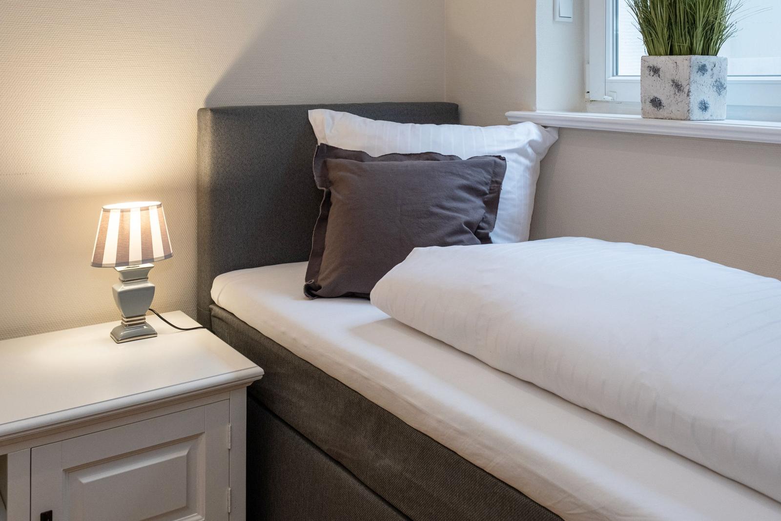 Ein Bett mit weißer Bettwäsche und einem Kissen. Neben dem Bett steht ein Nachttisch mit einer Lampe.