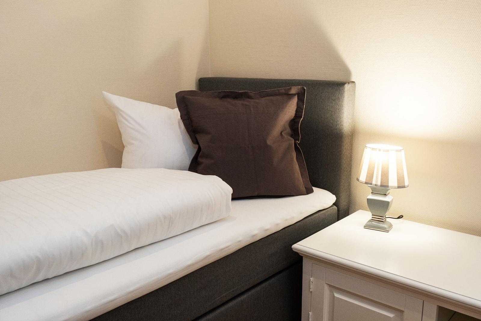 Ein Bett mit weißer Bettwäsche und einem braunen Kissen. Neben dem Bett steht ein Nachttisch mit einer Lampe.