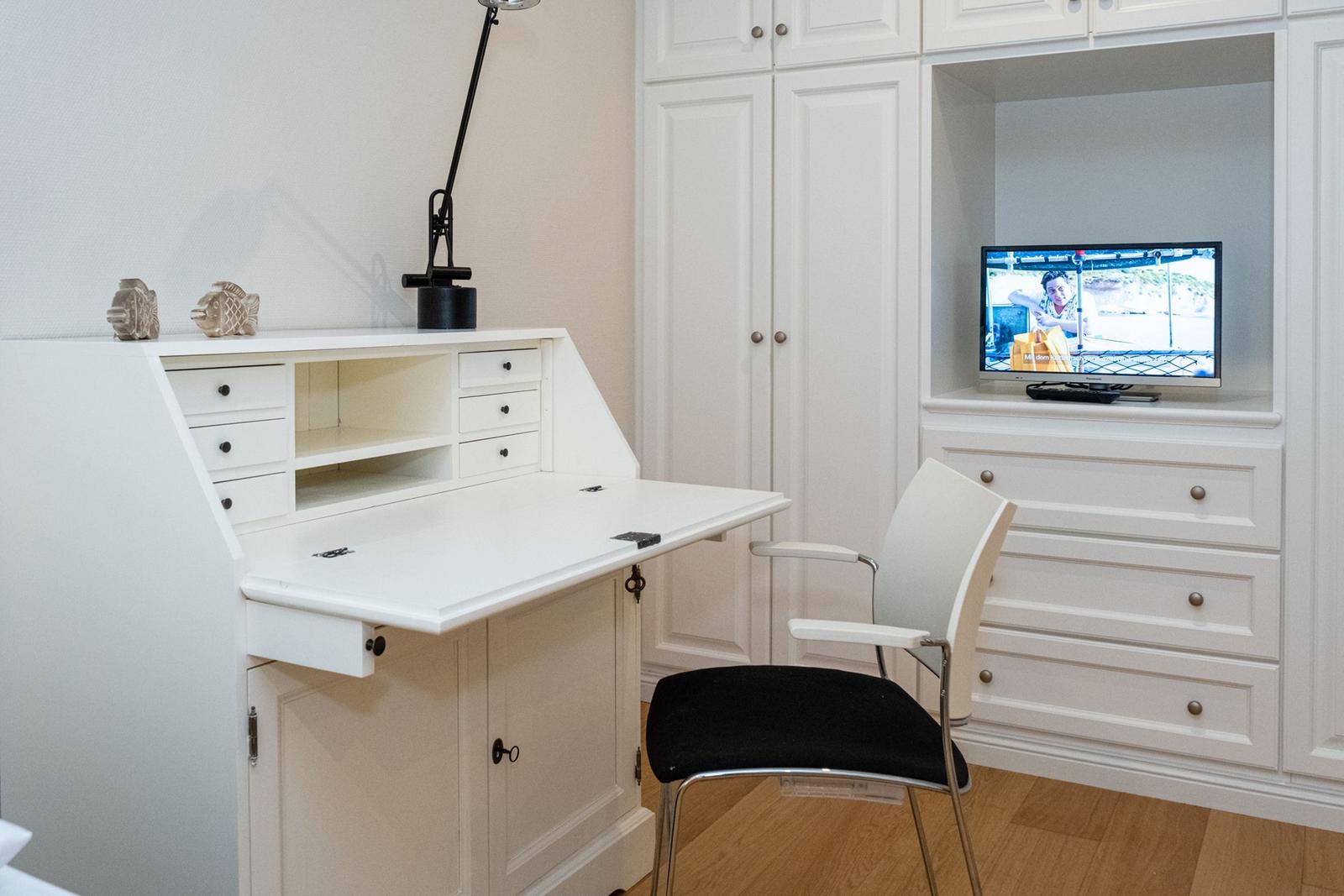 Arbeitsplatz mit Schreibtisch, Stuhl und Fernseher in weißer Einbauküche.