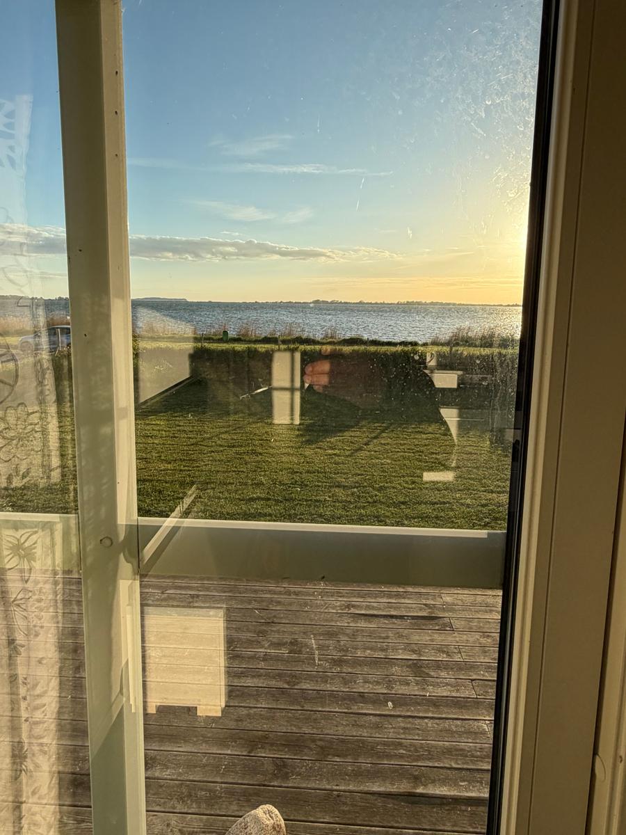 Blick auf einen See durch eine Glasschiebetür mit Holzterrasse und Rasenfläche.