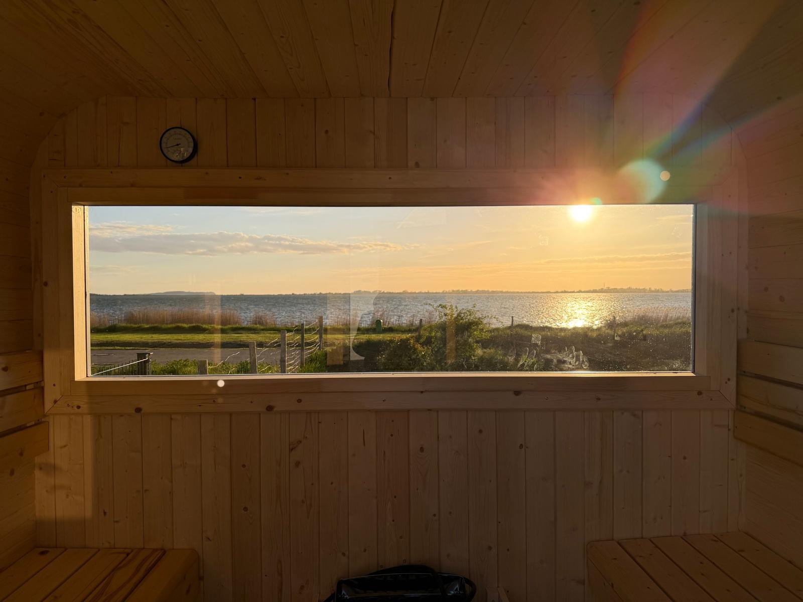 Sicht aus Holzsauna auf Sonnenuntergang über Wasser und Grünflächen