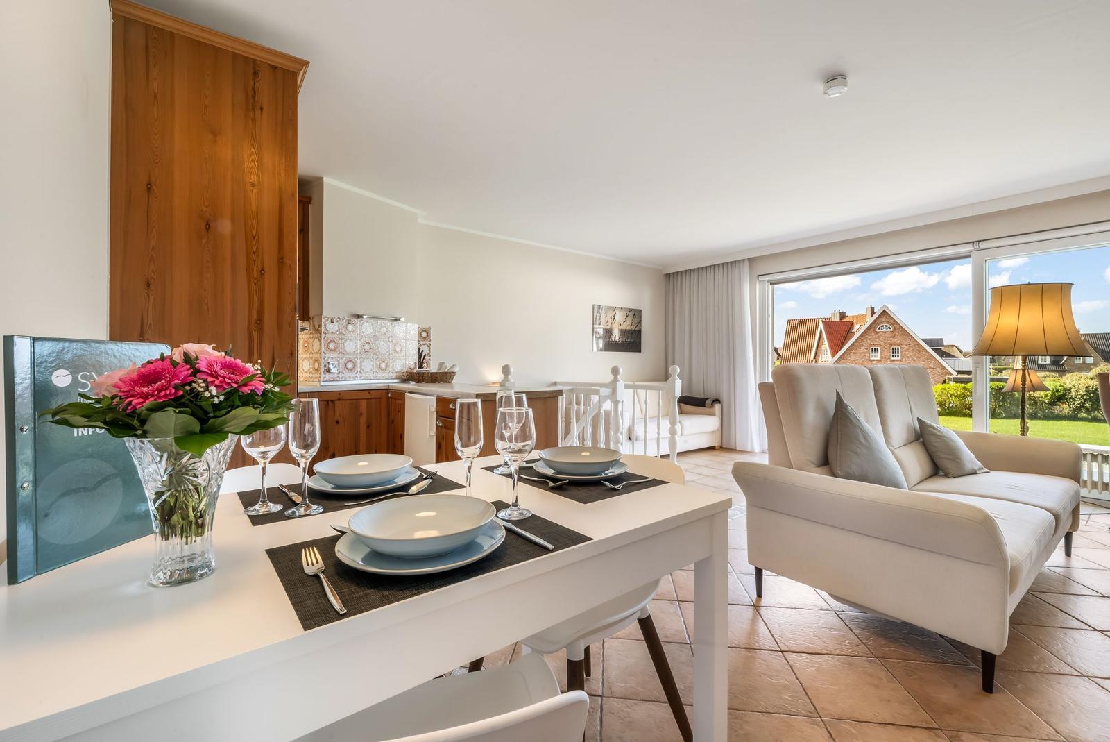Dining table with placemats, chairs, and flowers. Living area with sofa and garden view.