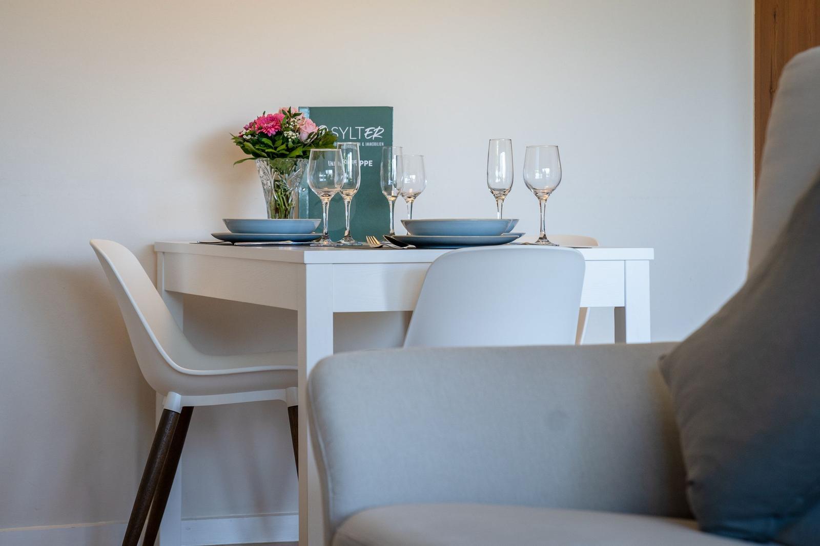 A white table with chairs, plates, and glasses. Flowers and a book are on it.