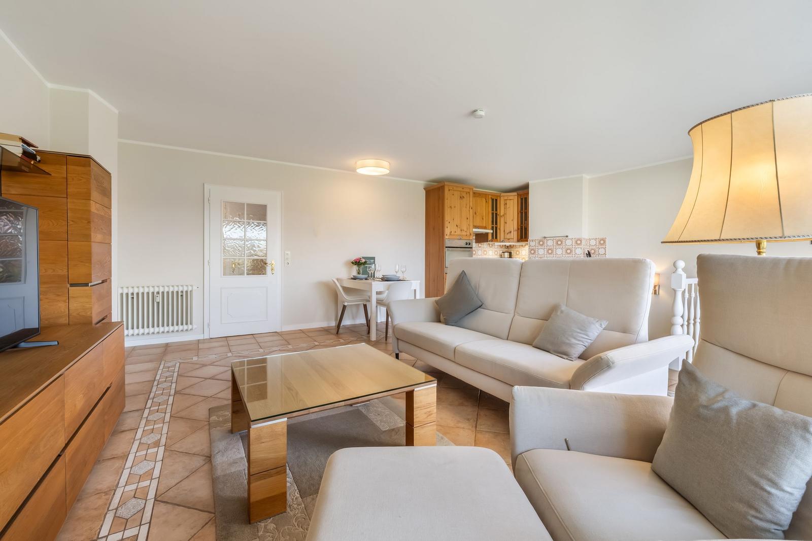 Living area with sofa, armchair, glass table, and kitchen area in the background.