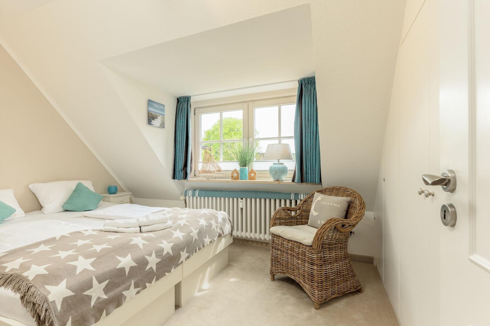 Ein Schlafzimmer mit Dachschräge, Bett, Stuhl und Fenster mit Vorhängen.