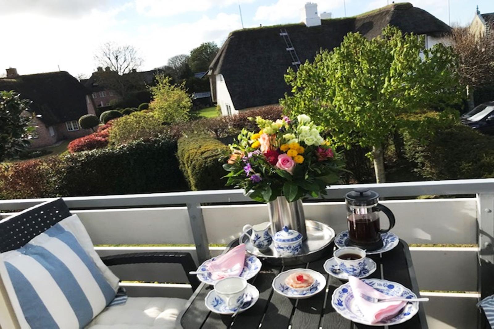 Terrasse mit Tisch, Stühlen und Blumenstrauß. Blick auf grüne Hecken und Häuser mit Strohdächern.