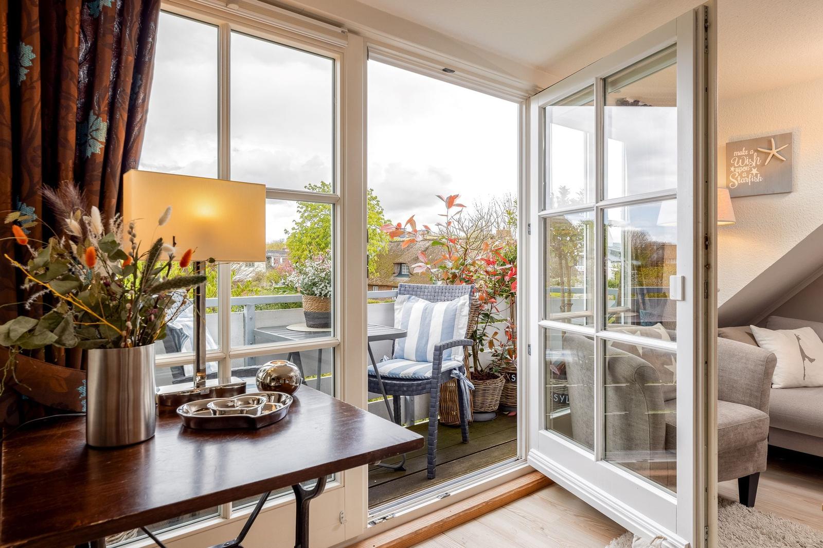 Terrasse mit Stuhl und Pflanzen. Blick ins Wohnzimmer mit Sofa und Wandbild.