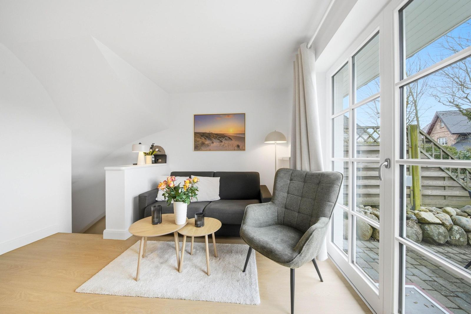 Living room with sofa, armchair, and table. Large window view to outdoor terrace.