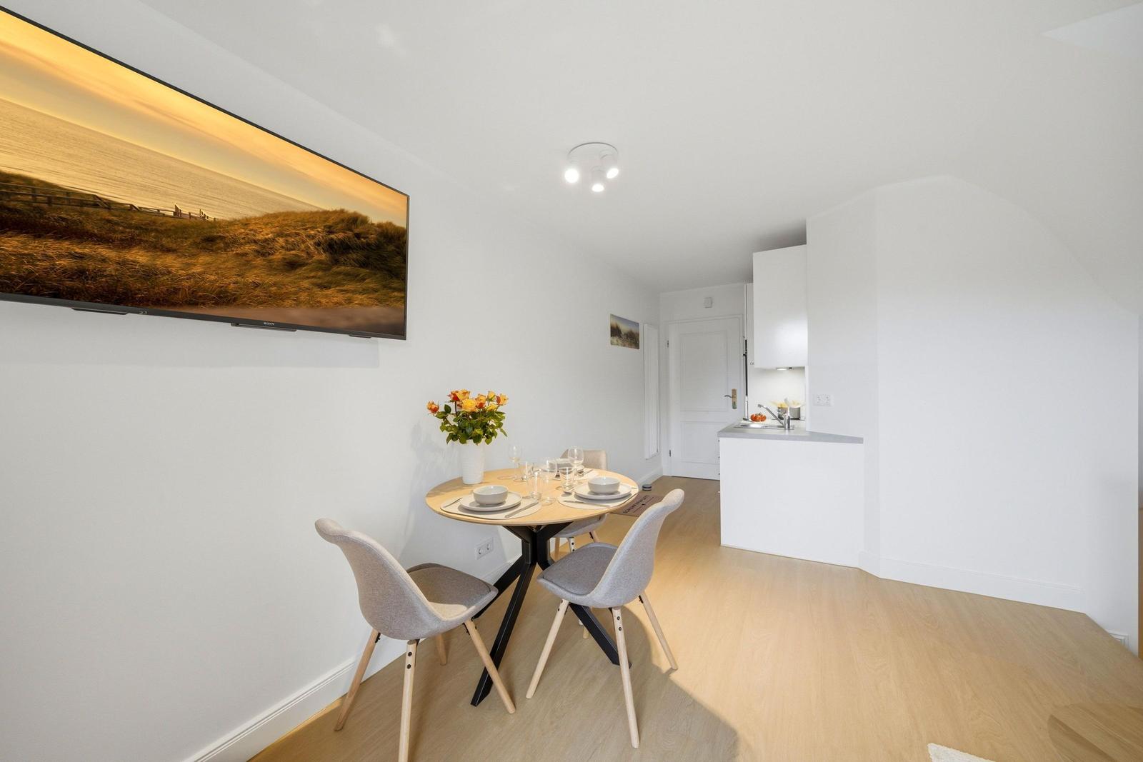 Dining area with table and chairs, kitchen in background, wall art and wooden floor.