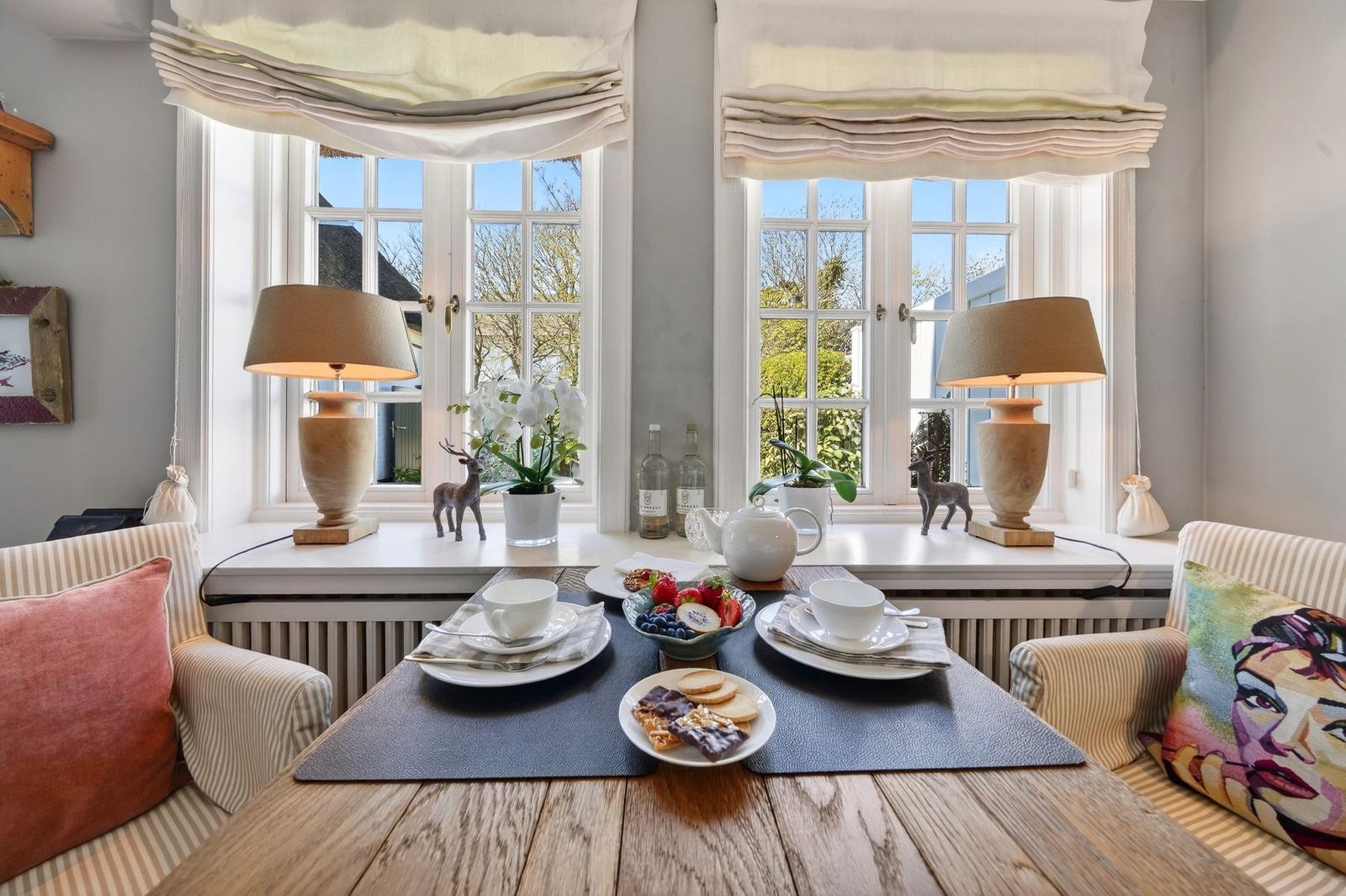 Tisch mit Frühstück vor zwei Fenstern mit Lampen und Pflanzen.
