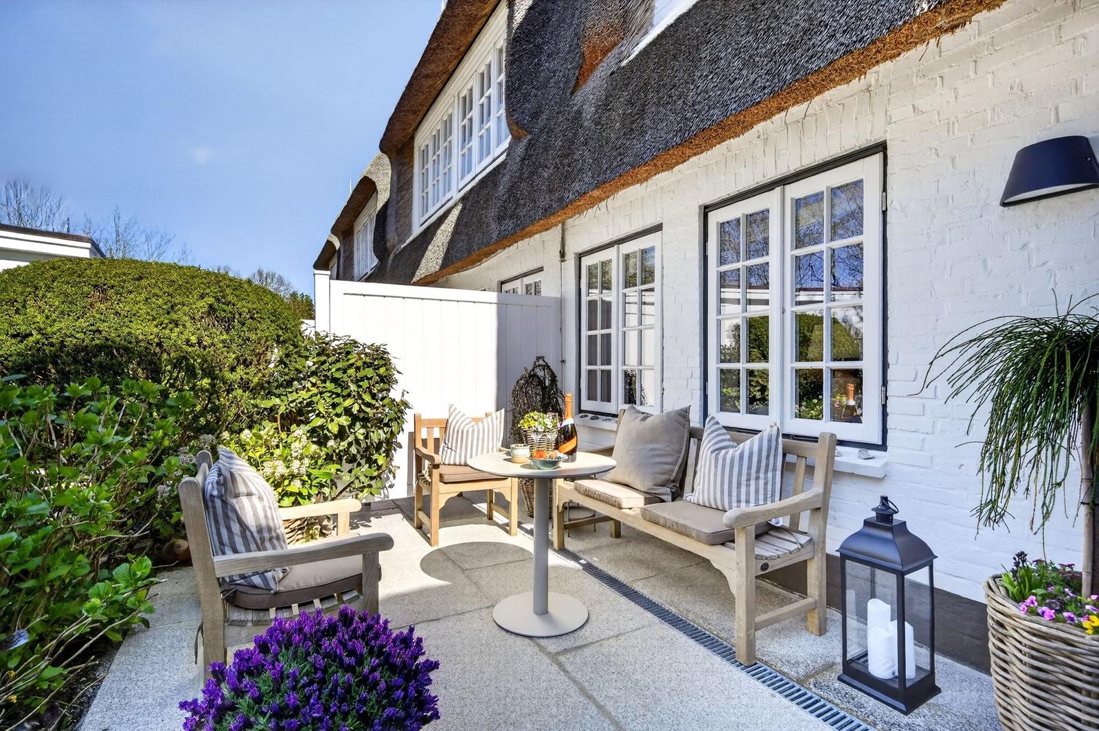 Terrasse mit Sitzgelegenheiten, Tisch und Blumen vor einem Haus mit Strohdach und weißen Wänden.