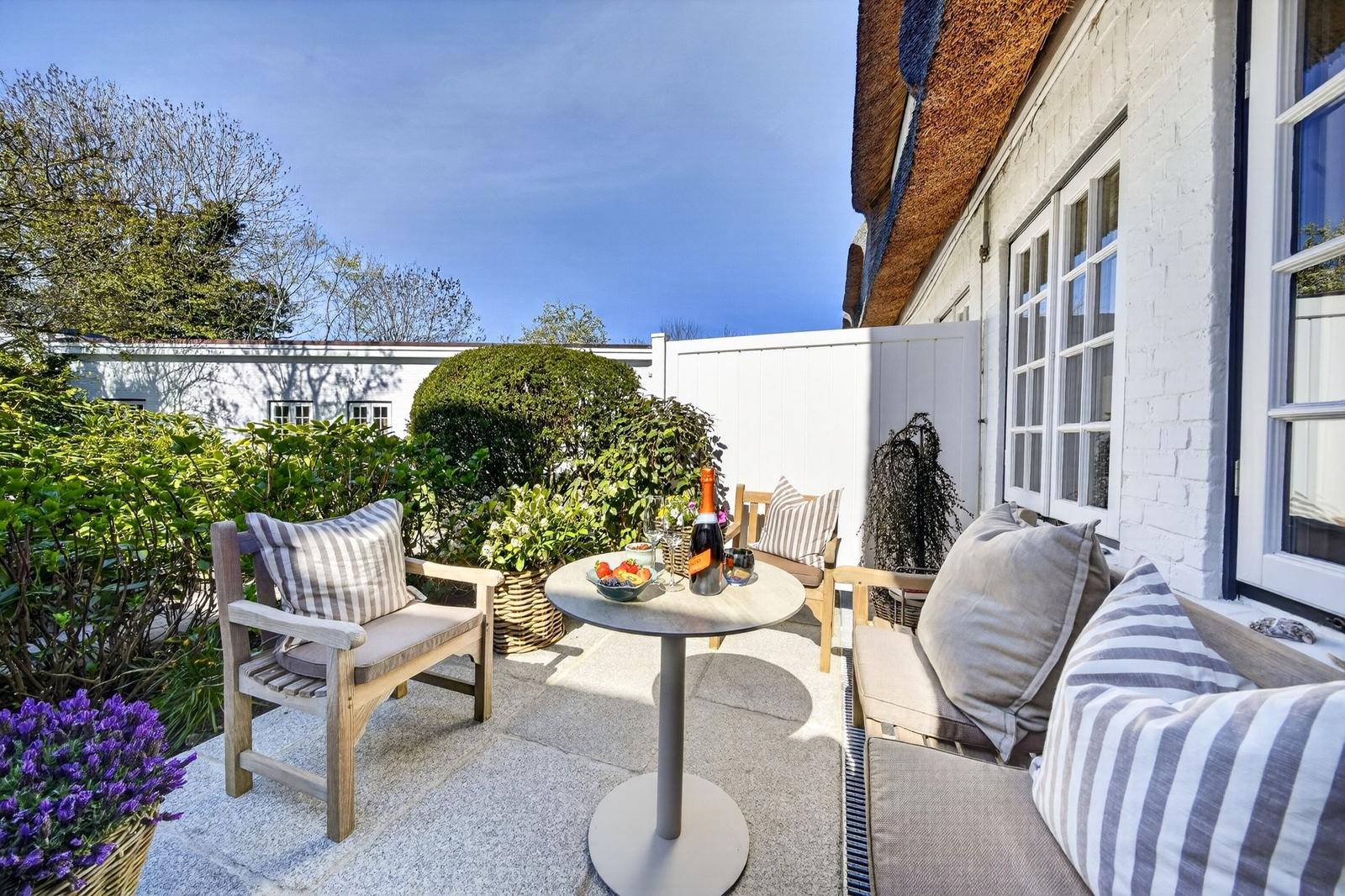 Terrasse mit Tisch, Stühlen und Blumen. Hintergrund: weißer Zaun und Hauswand.