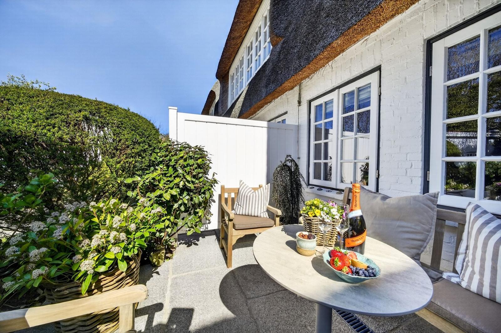 Terrasse mit Tisch, Stühlen und Pflanzen vor weißem Haus mit Dachschindeln.