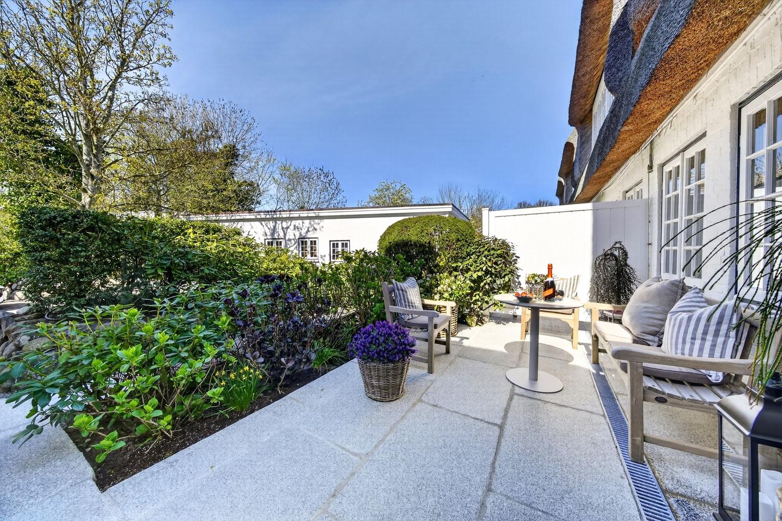 Terrasse mit Sitzgelegenheiten, Pflanzen und Tisch. Hinterhof mit Hecke und weißem Gebäude.
