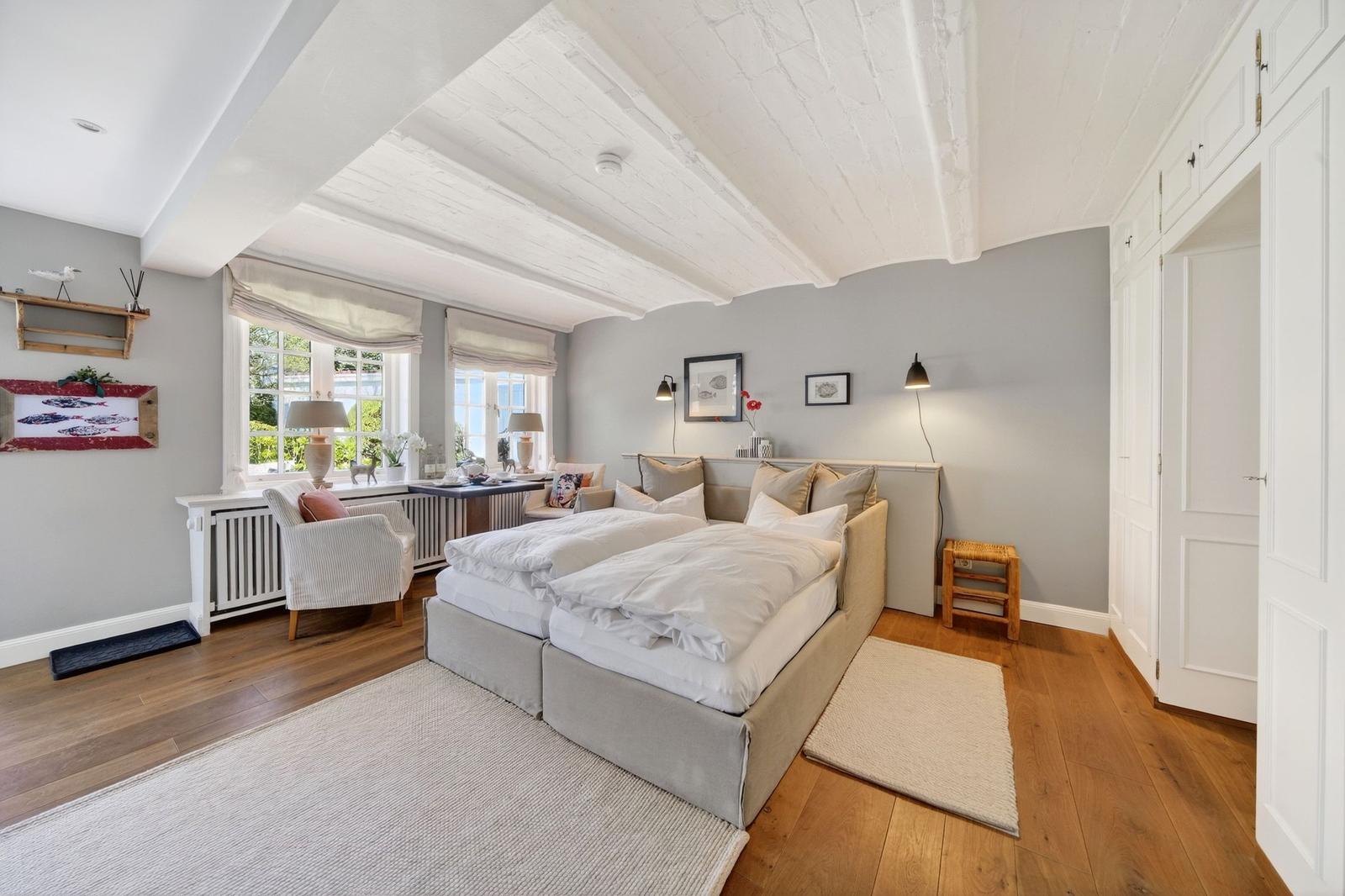 Schlafzimmer mit Doppelbett, Holzboden und weißen Balkendecken. Zwei Fenster mit Blick nach draußen.