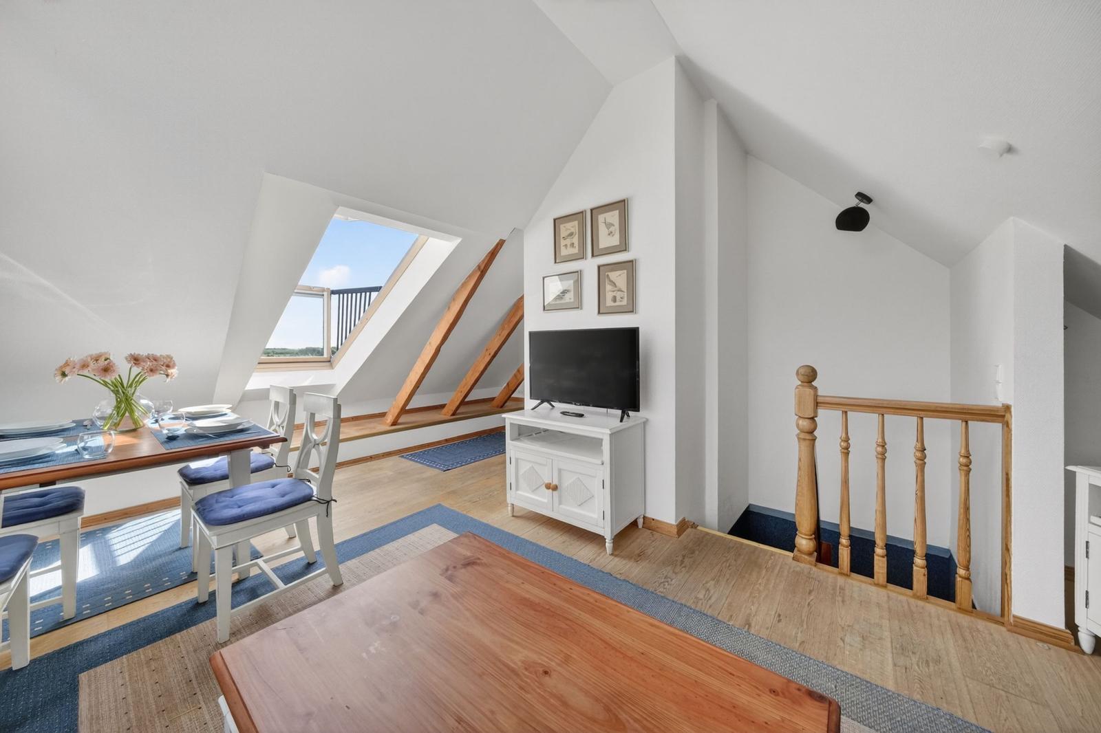 Dachgeschosswohnung mit Essbereich, TV und Balkonzugang. Holzdielen und weiße Wände.