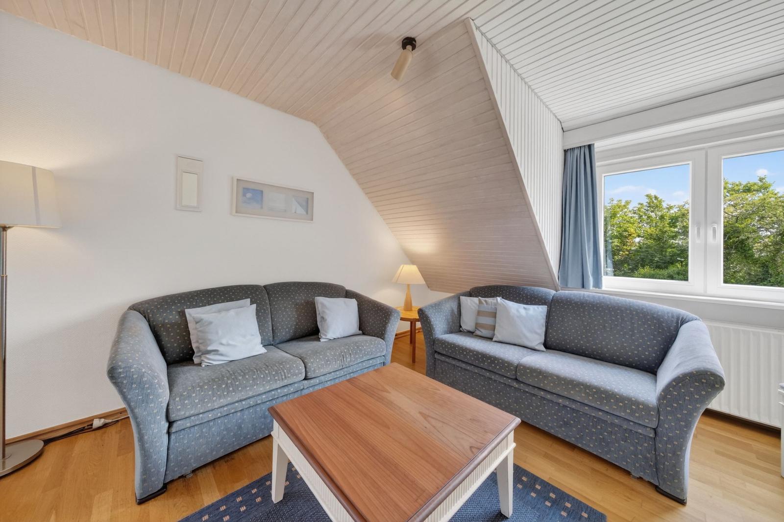 Zwei Sofas mit Kissen stehen um einen Holztisch herum. Fenster zeigt grüne Bäume. Holzdecke und Boden.