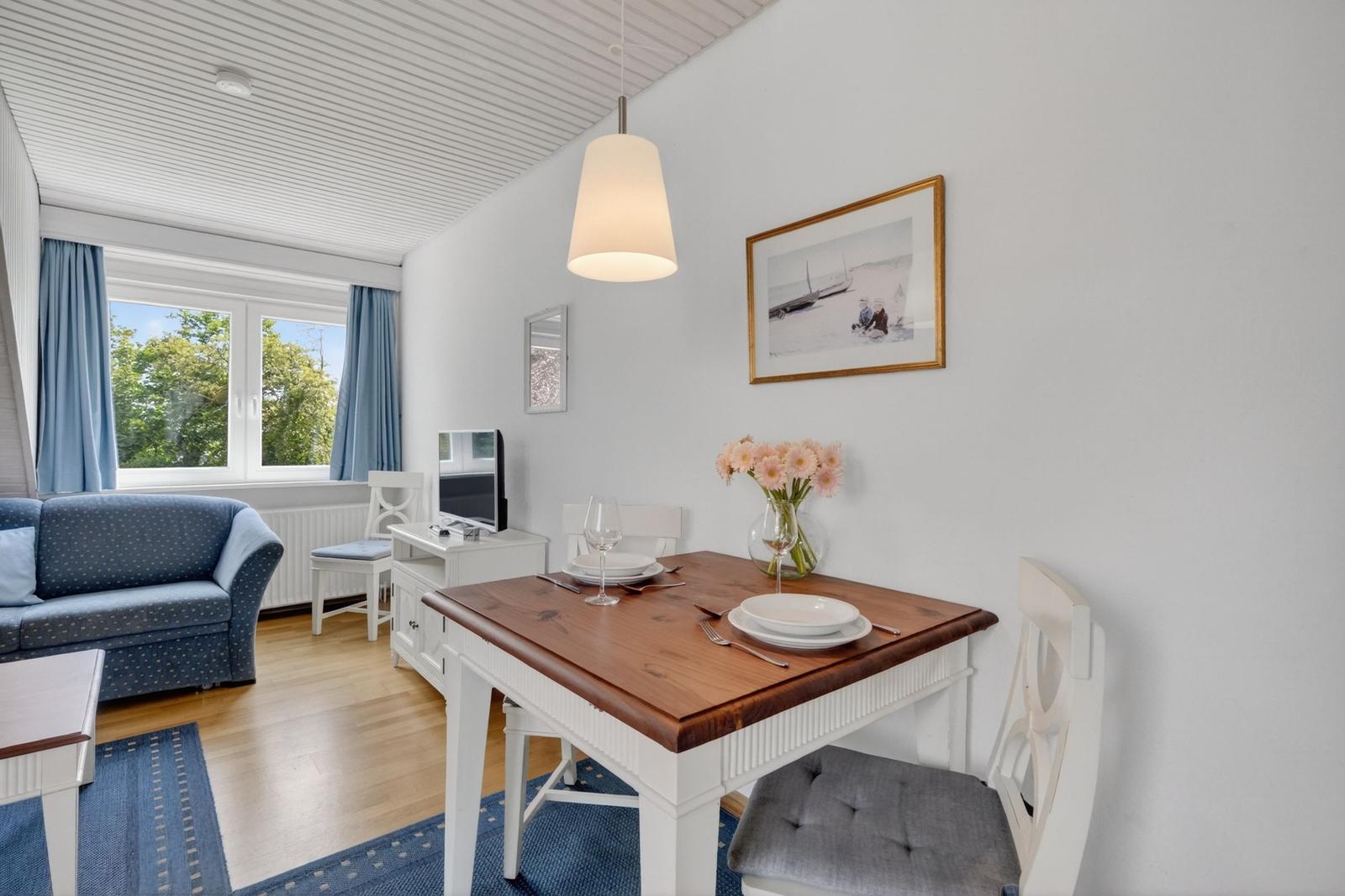 Essbereich mit Holztisch, Stühlen und Blumen. Wohnbereich mit Sofa und Fenster.