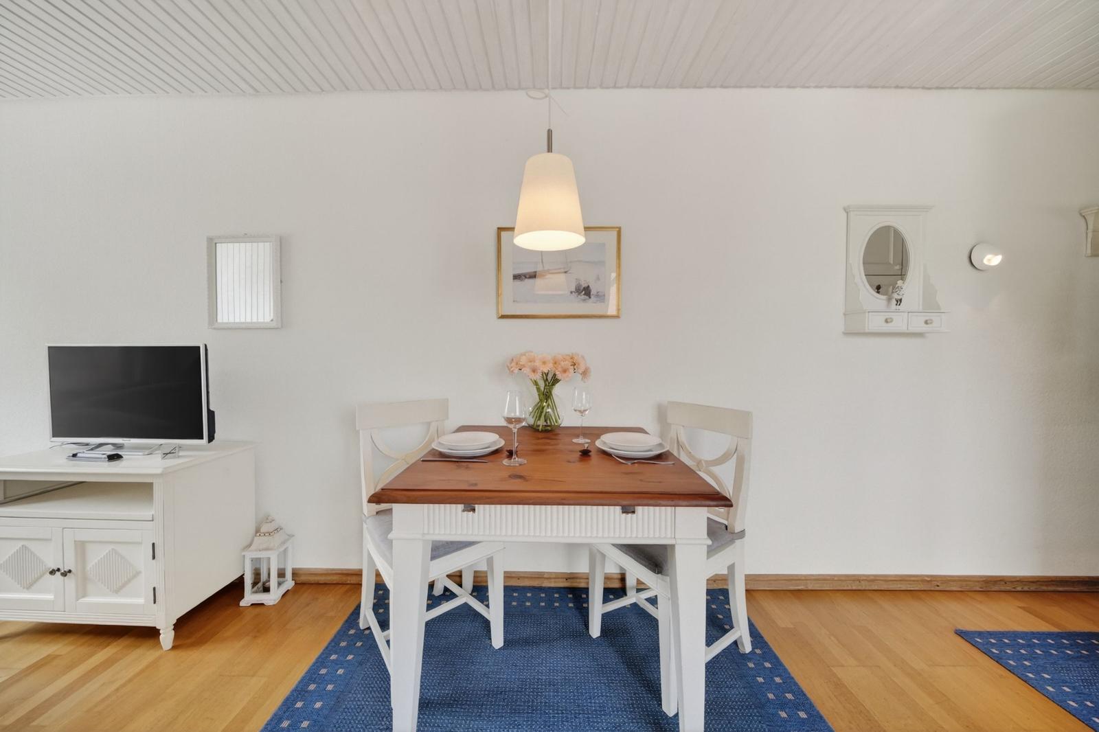 Esszimmer mit Holztisch, weißen Stühlen und blauem Teppich. An der Wand hängt ein Bild und ein Spiegel.
