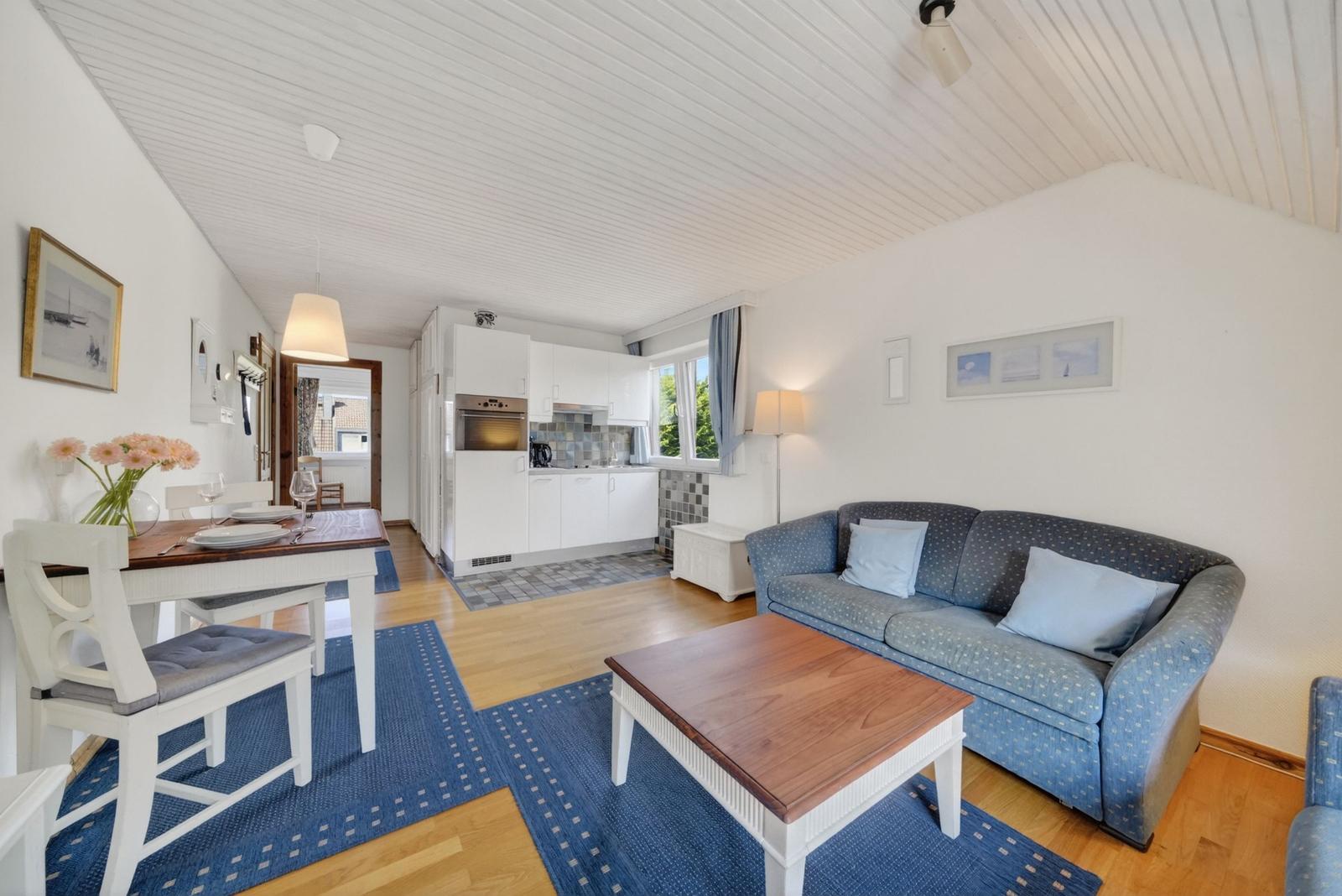 Wohn- und Essbereich mit Sofa, Tisch und Küche. Holzboden und weiße Wände.