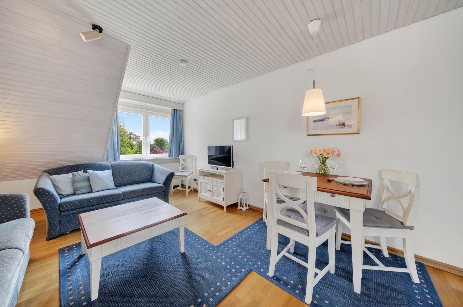 Wohn- und Essbereich mit Sofa, Tisch und Stühlen. Fenster mit Blick auf Grün.