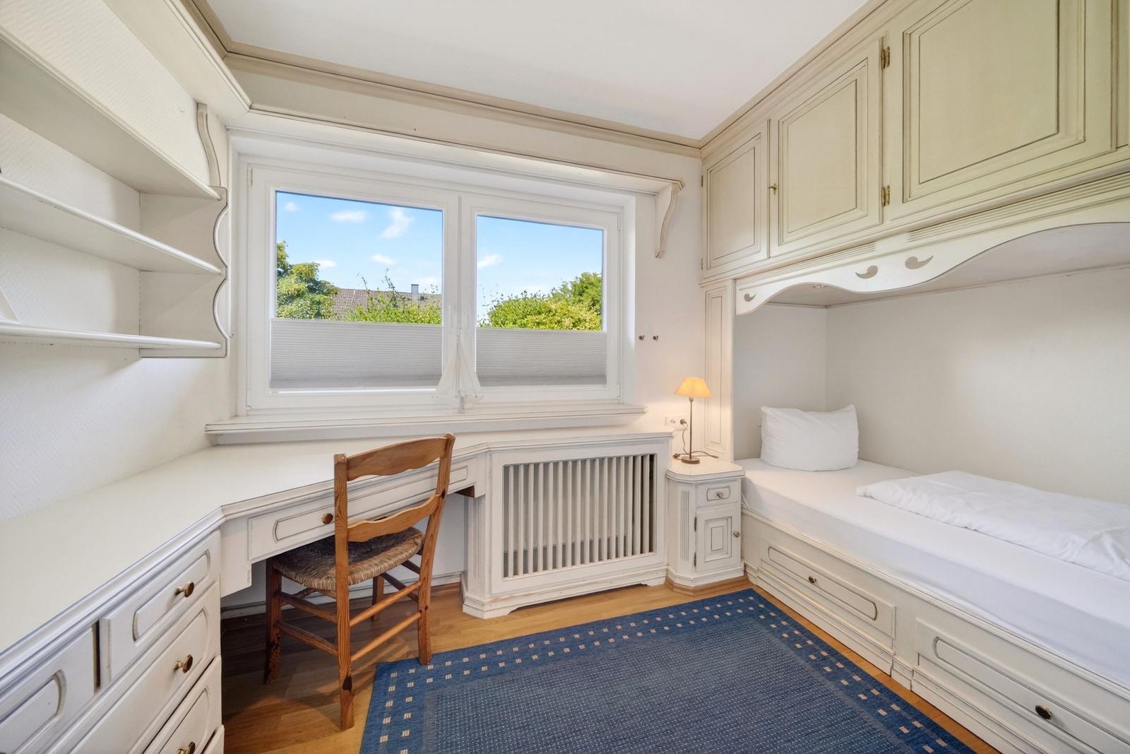 Schlafzimmer mit Schreibtisch, Bett und Fenster. Holzfußboden und blaues Teppichmuster.