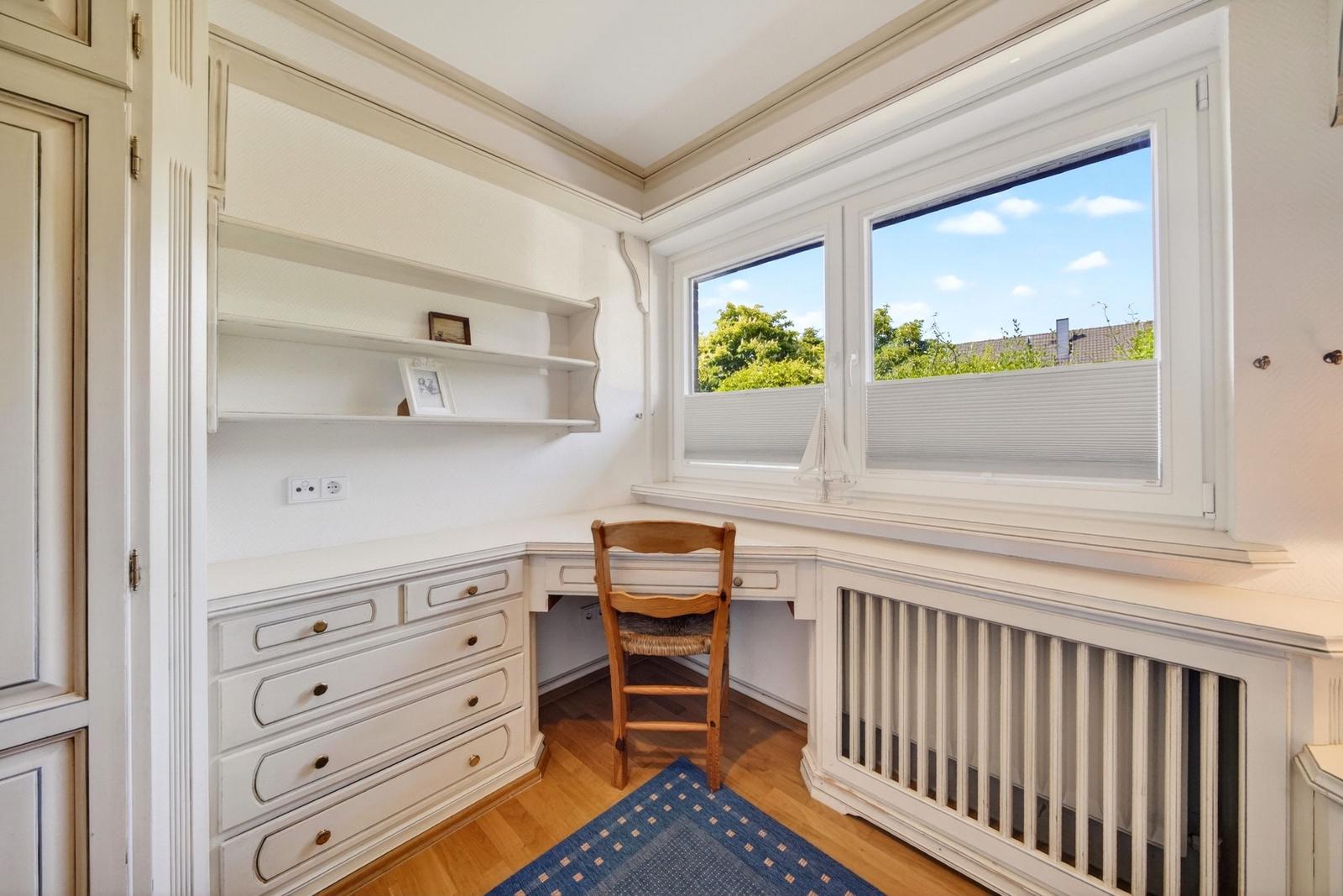 Ein Arbeitszimmer mit Schreibtisch, Stuhl und Fensterblick auf Bäume.