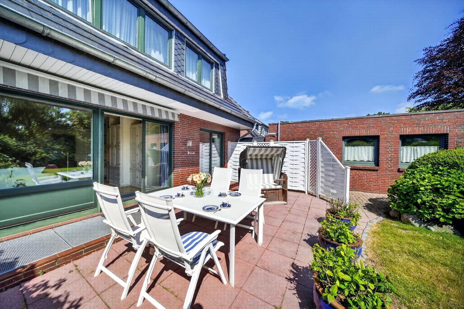 Terrasse mit Tisch, Stühlen und Liege. Gartenhaus und Pflanzen im Hintergrund.