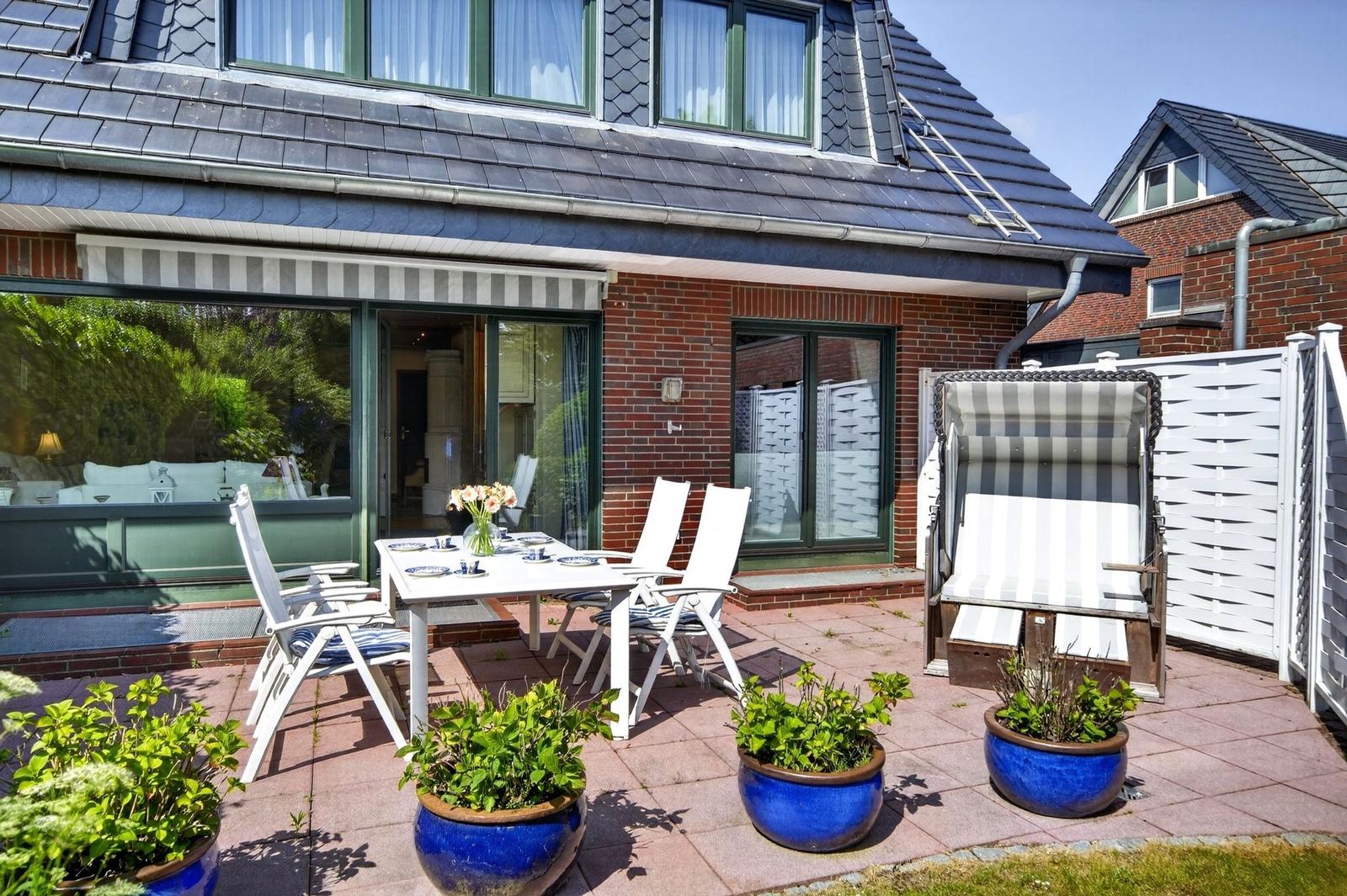 Terrasse mit weißem Tisch, Stühlen und Strandkorb. Blaue Töpfe mit Pflanzen. Haus mit Glasfront und Ziegelfassade.