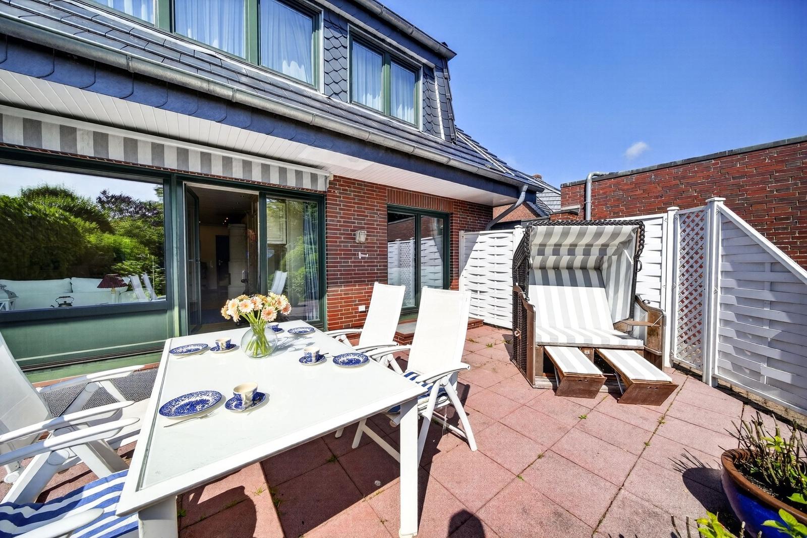 Terrasse mit Tisch, Stühlen und Strandkorb. Haus mit Glasfront und rotem Ziegelmauerwerk.