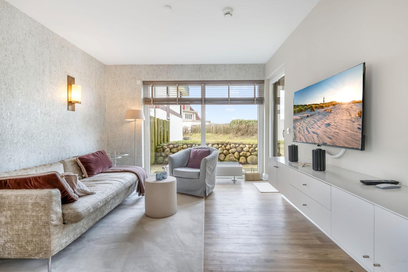 Wohnzimmer mit Sofa, Sessel und Fernseher an der Wand. Durchgang zum Garten.