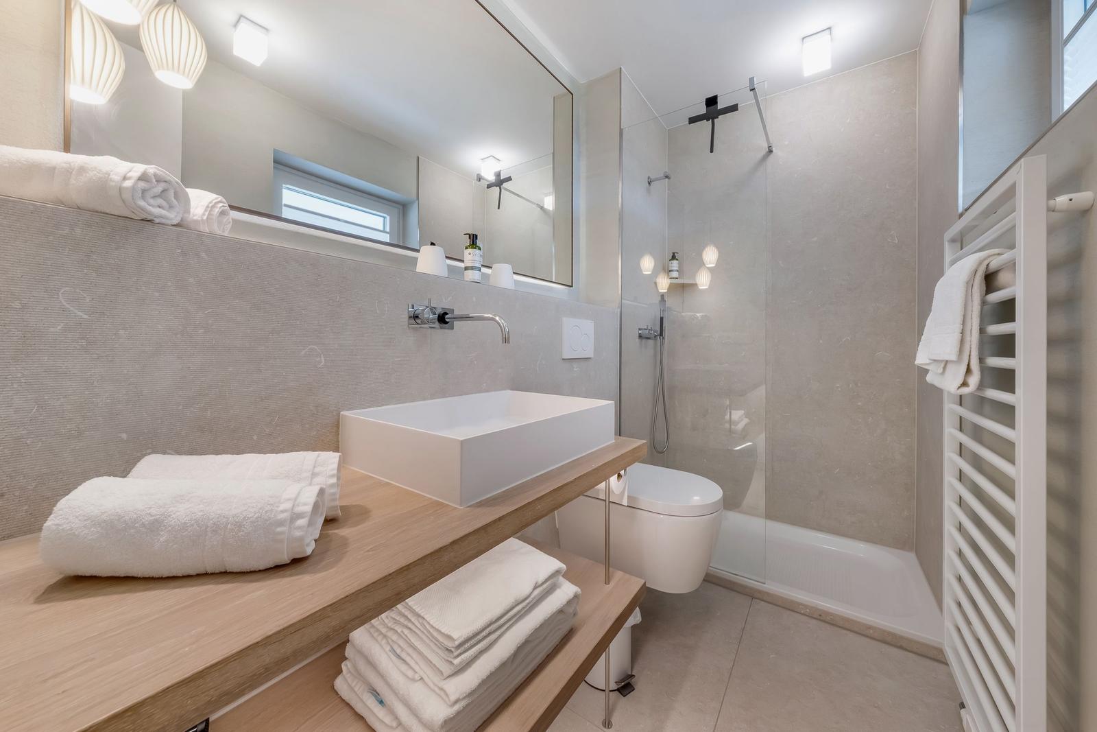 Badezimmer mit Dusche, Waschbecken und Toilette. Weiße Handtücher auf Holzregal.