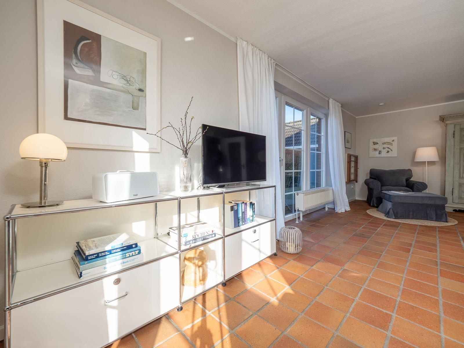 Wohnzimmer mit TV, Bücherregal und Terrassentür. Terrakottaboden und graue Sitzgelegenheit.
