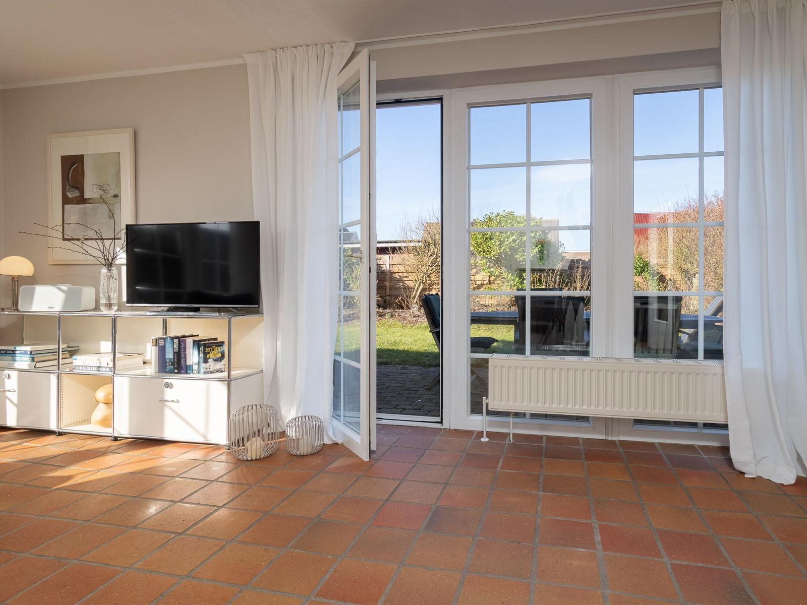 Wohnzimmer mit Terrassentür, TV und Terrasse. Terrakottafußboden und weißer Heizkörper.