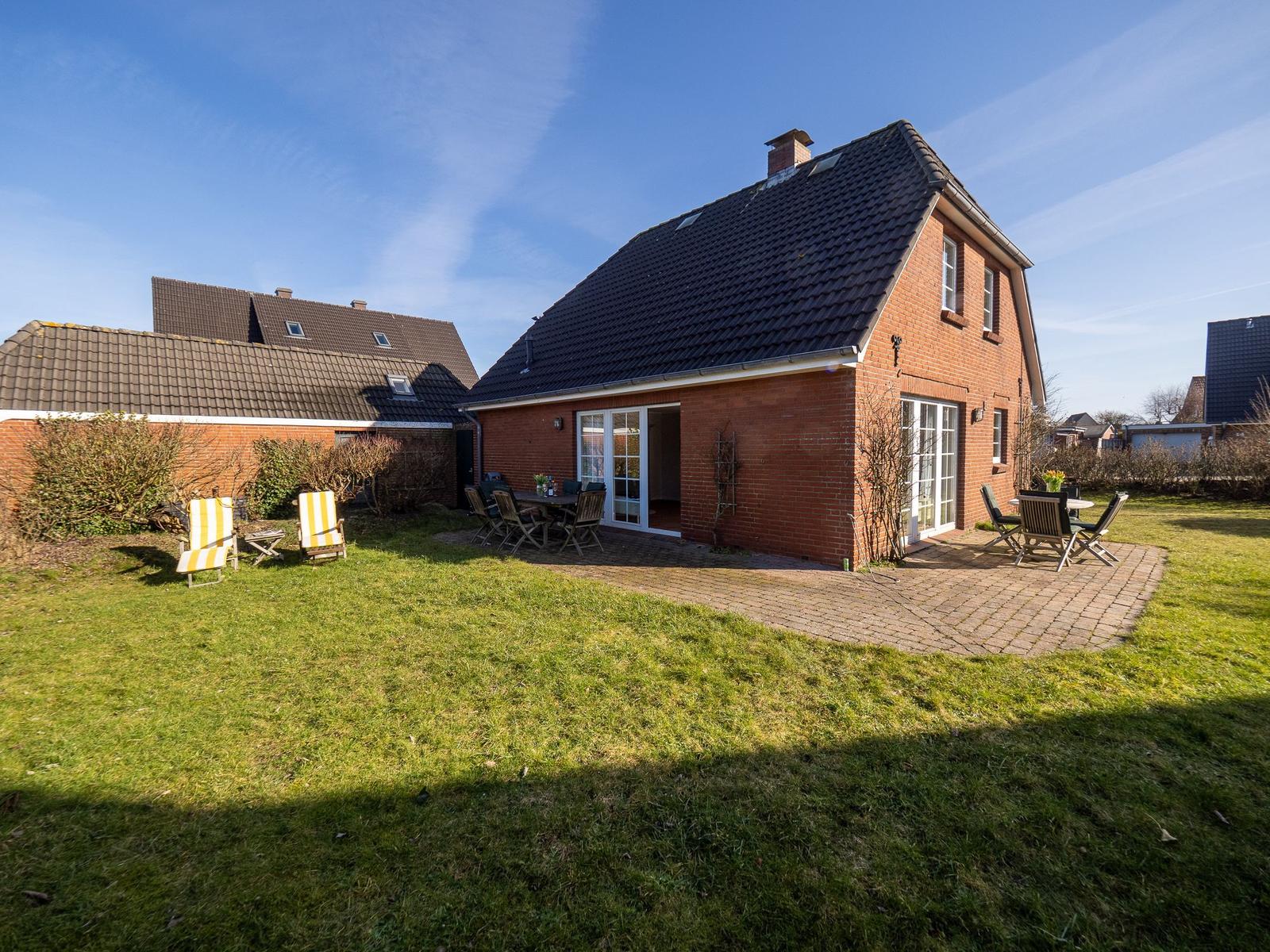 Rotes Backsteinhaus mit Garten, Terrasse und Gartenmöbeln unter blauem Himmel.