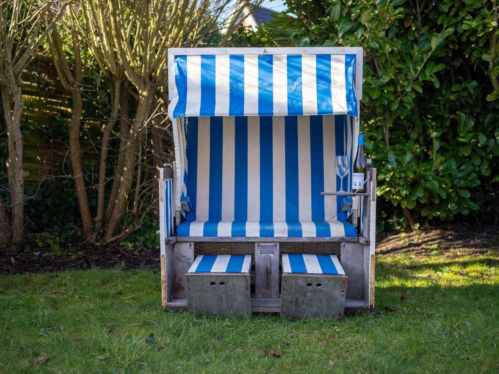 Ein blau-weiß gestreifter Strandkorb steht auf einem Rasen. Auf dem Tisch steht ein Glas und eine Flasche.
