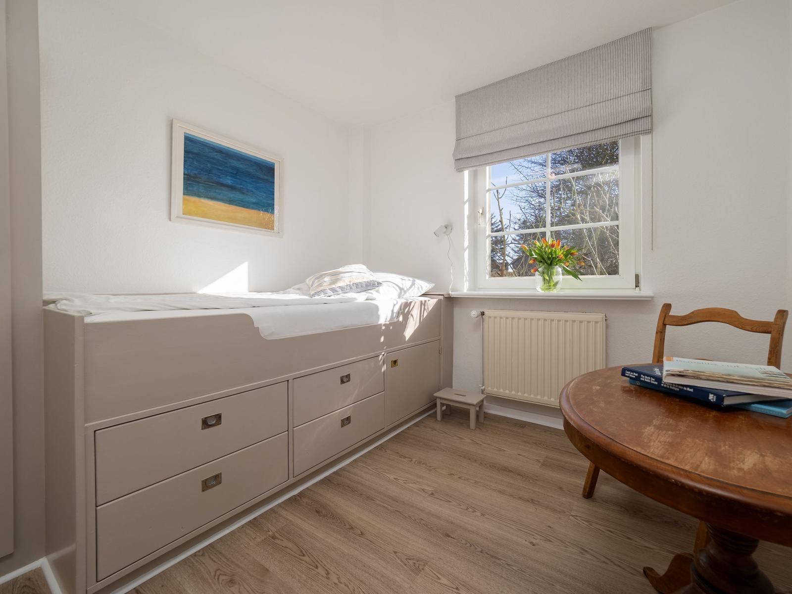 Schlafzimmer mit Bett, Schubladen, Tisch und Stuhl. Fenster mit Blumen und Bild an der Wand.