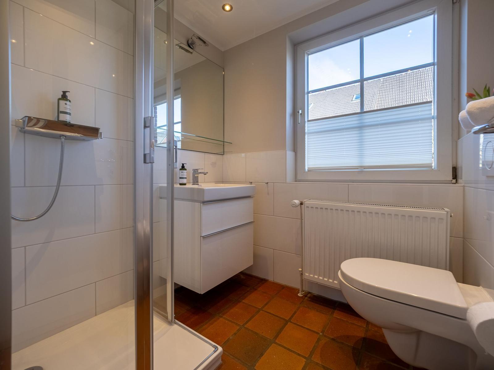 Badezimmer mit Dusche, Waschbecken und Toilette. Fenster mit Jalousie und Heizkörper.