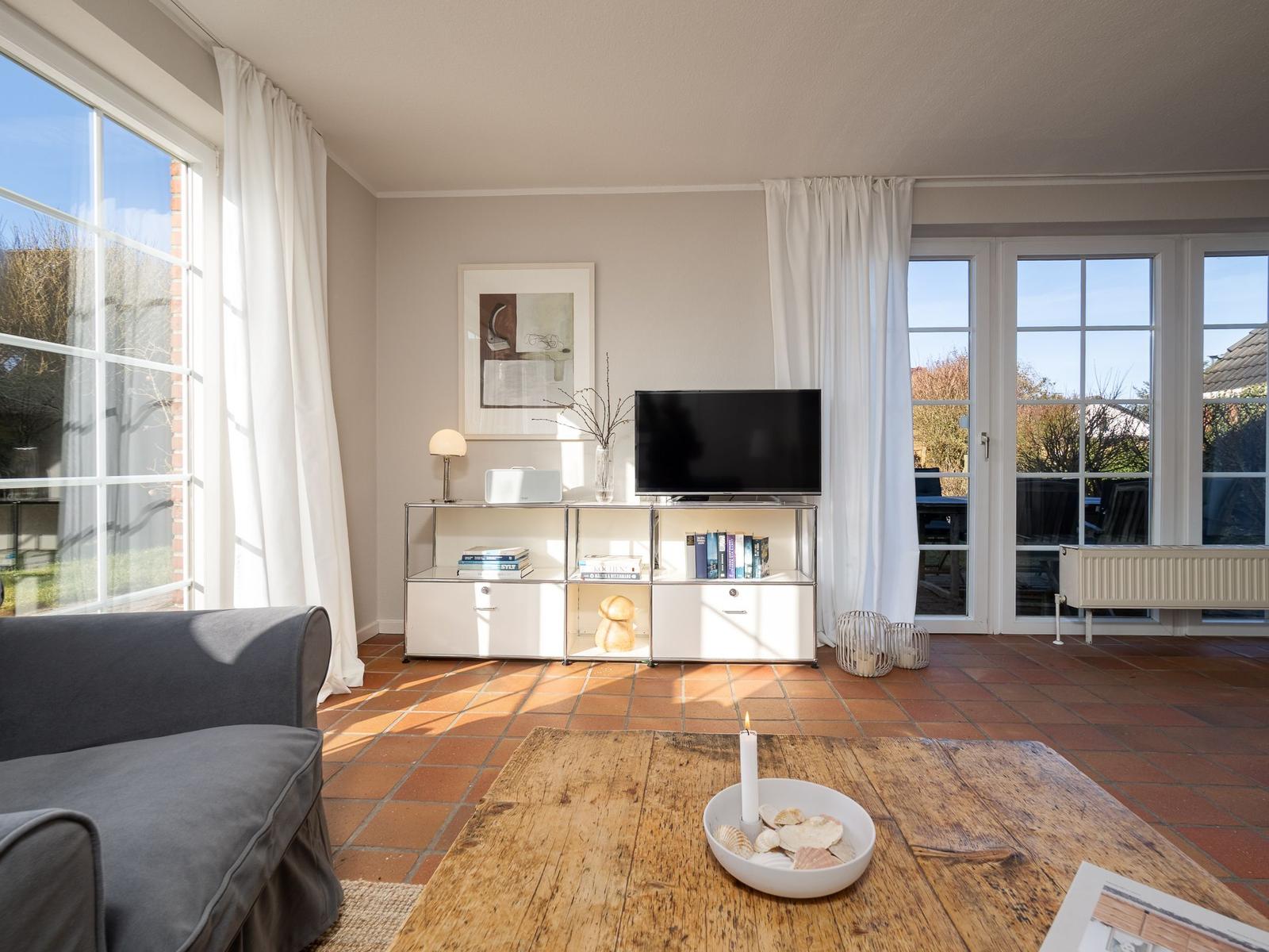 Wohnzimmer mit TV, Holztisch und großen Fenstern. Terrakottaboden und weiße Vorhänge.