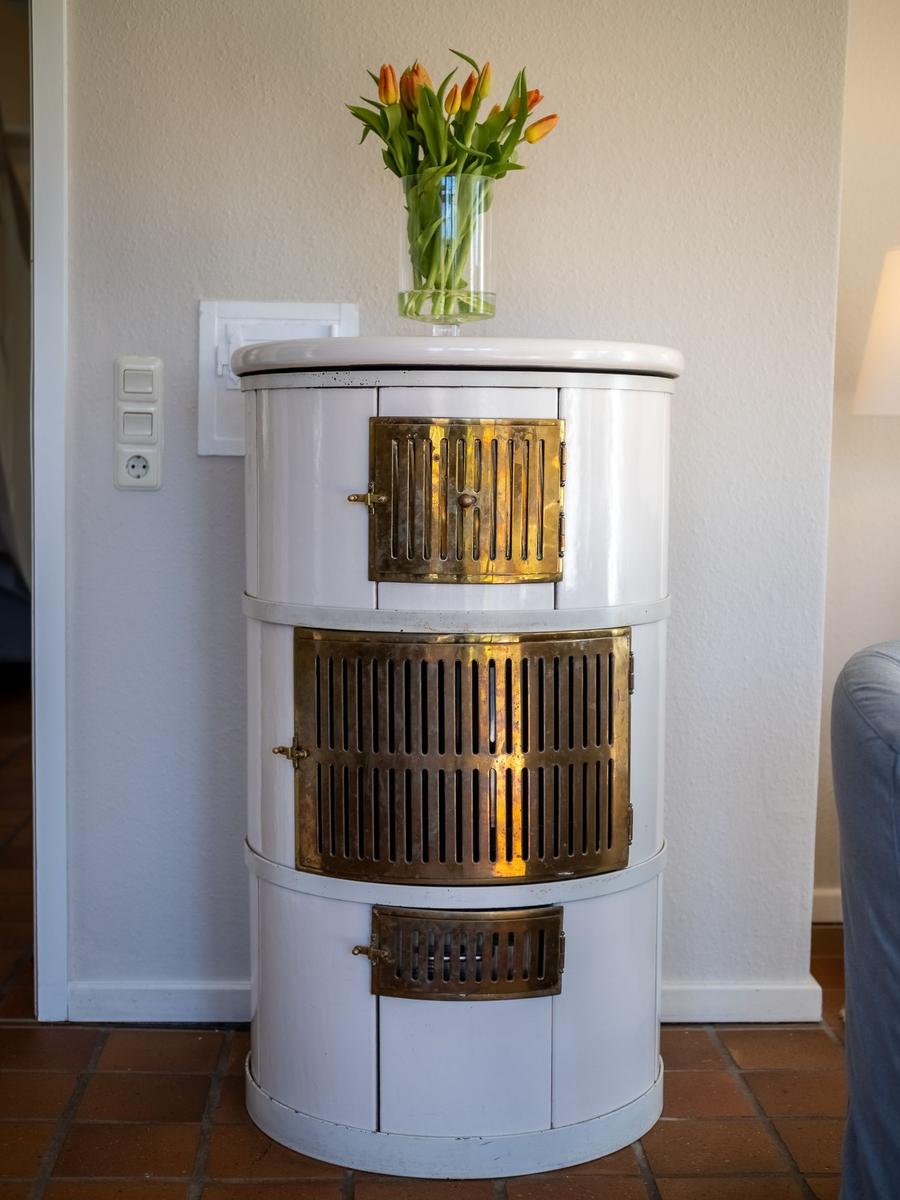 Ein weißer, zylindrischer Ofen mit goldenen Gittertüren und einer Tulpenvase darauf.