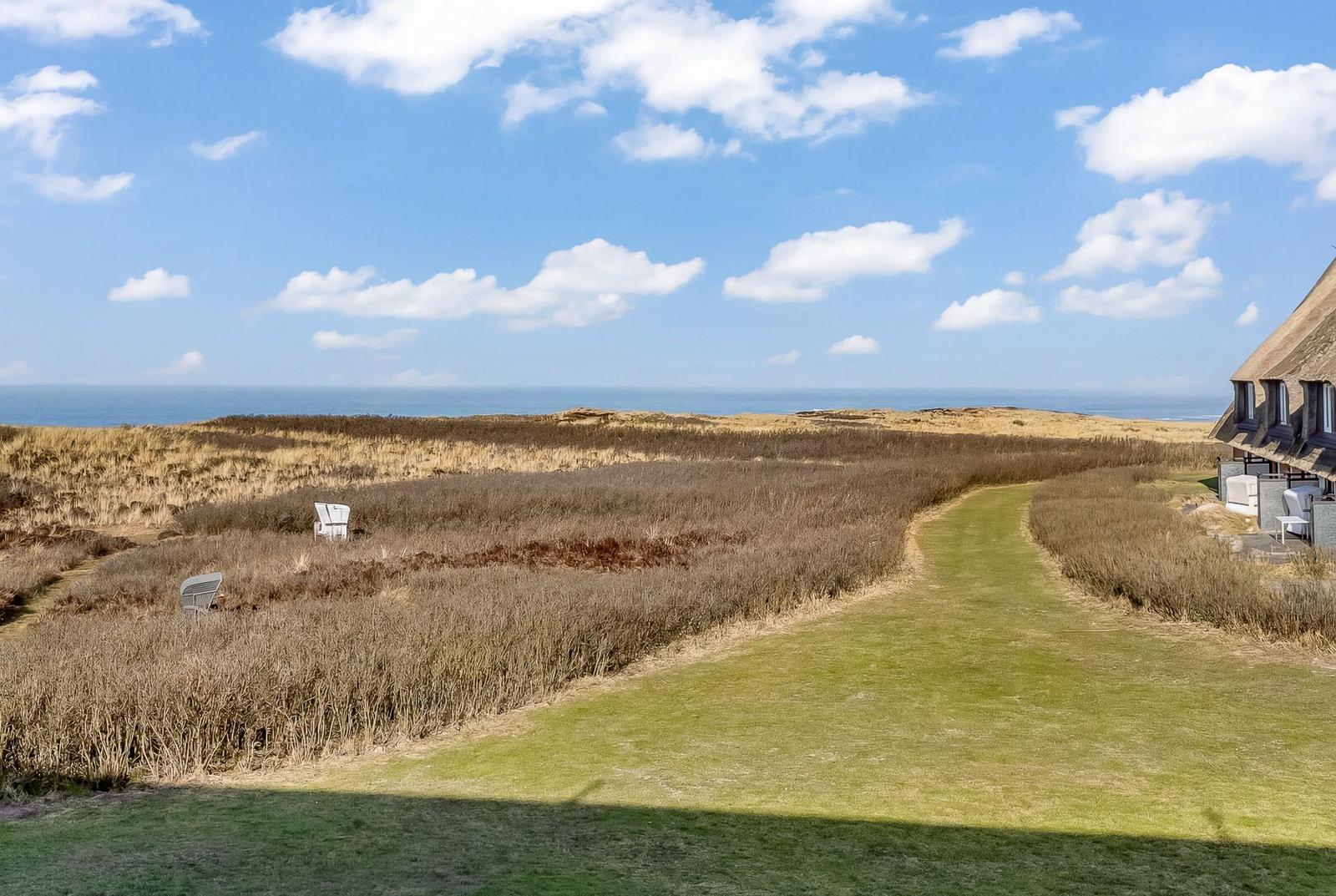 Green path leads through dry grass to the beach promenade and a beach house.