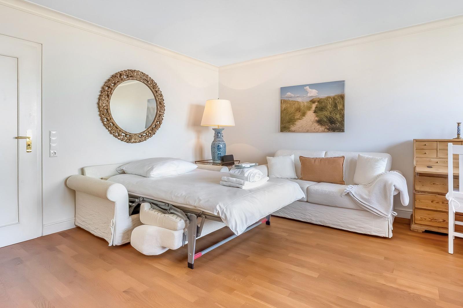 Bedroom with sofa, bed, and wooden floor. Wall art and mirror visible.