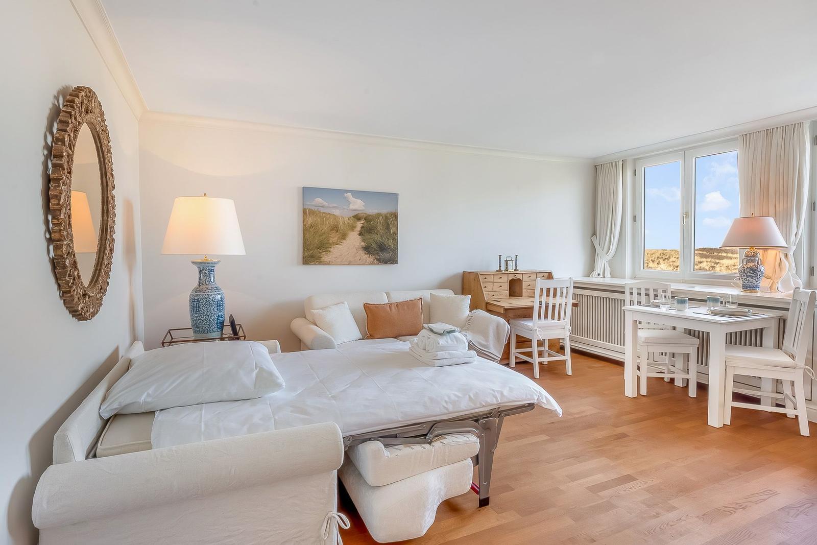 A bedroom with a white bed, sofa, table, and chairs. Large windows with a view of a meadow.