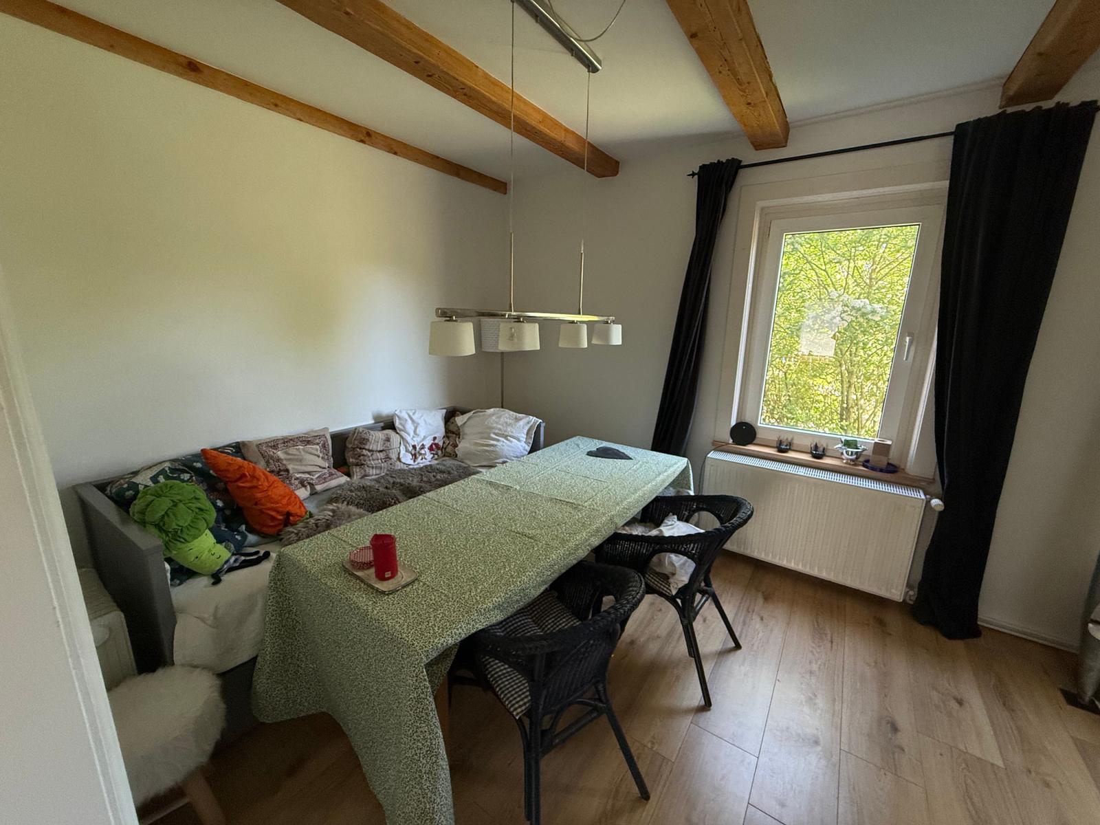 Ein Raum mit Tisch, Stühlen und Sofa. Holzbalken an der Decke und Fenster mit Blick auf Bäume.